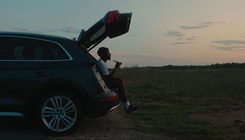 Person sitting in open car trunk at dusk in an open field, gazing toward the horizon.