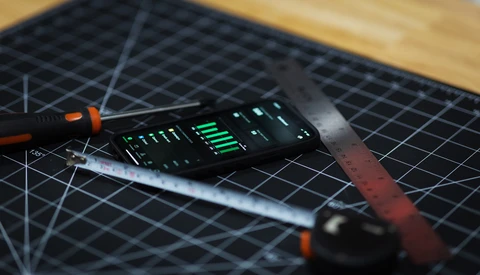 Electronic device with illuminated green LED display sits on a black cutting mat next to precision tools.