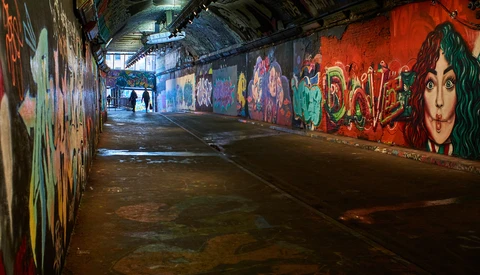 Underground tunnel lined with colorful graffiti murals under industrial lighting.