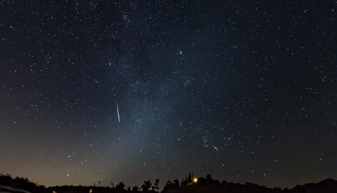 Meteor streaking across starry night sky above silhouetted landscape with warm lights below.