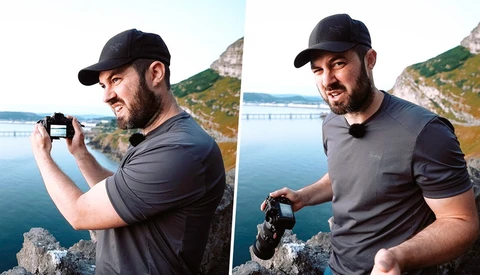 Photographer holding a compact camera while standing on a rocky cliff overlooking a calm blue lake and forested shoreline.