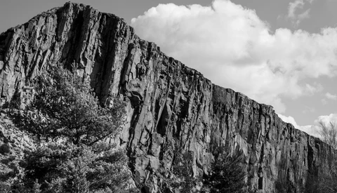 Dramatic columnar basalt cliff face rises against a cloudy sky, with vegetation at its base.