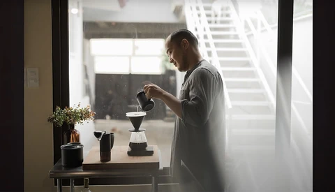 Man in casual shirt preparing pour-over coffee near a bright window with natural morning light.