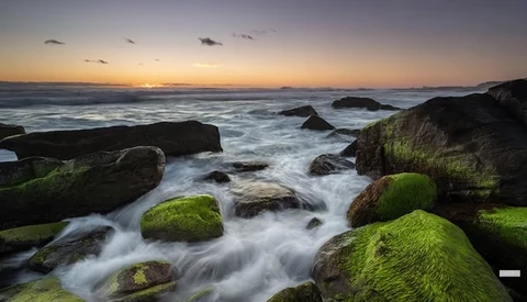 Coastal landscape at sunset with moss-covered rocks and flowing water in foreground.