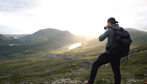 Photographer with camera and backpack capturing landscape from hilltop at golden hour.