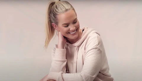 Young woman with blonde hair in a cream-colored hoodie, smiling with hand near face against neutral background.