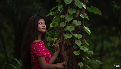 Woman in red polka dot top posing against ivy-covered tree trunk in forest setting.