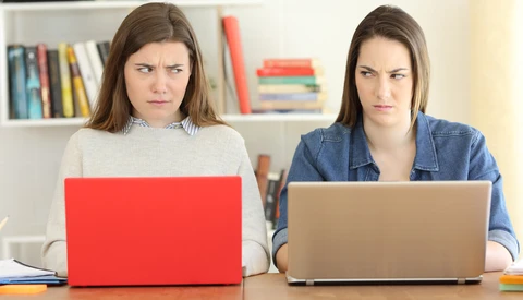 Two women sitting at a desk with laptops, both looking directly at the camera with serious expressions.