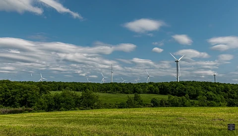 Wind turbines dotting a forested landscape under a partly cloudy sky.