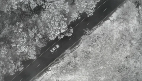 Aerial view of a vehicle driving on a dark asphalt road cutting through snowy forest terrain.
