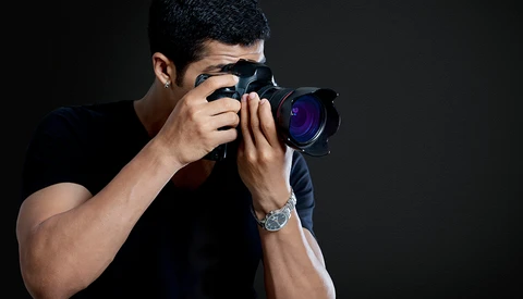Male photographer holding DSLR camera up to his face, shooting against dark background.