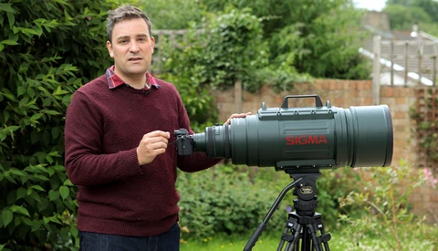 Man holding a Sigma telephoto lens mounted on a tripod in a garden setting.