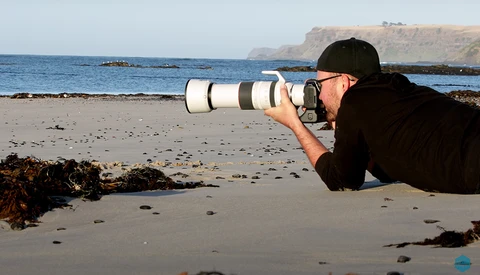 Photographer lying on beach sand with telephoto lens pointed toward ocean and distant cliffs.