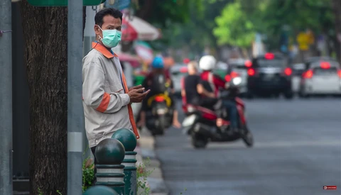 Man wearing surgical mask and orange safety vest standing against pole on urban street.
