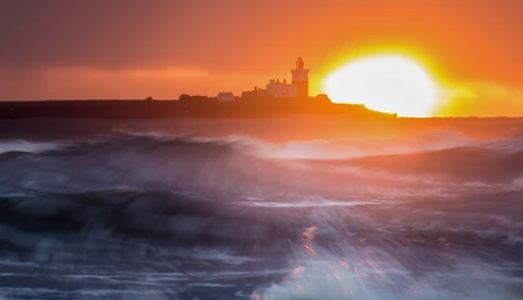 Coastal city skyline silhouetted against a bright sun setting over turbulent ocean waves.