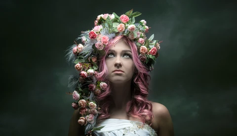 Woman with long pink hair wearing an elaborate floral crown of pink and white roses against a dark green background.