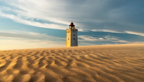 Abandoned yellow lighthouse tower standing alone in vast desert sand dunes under dramatic sky.