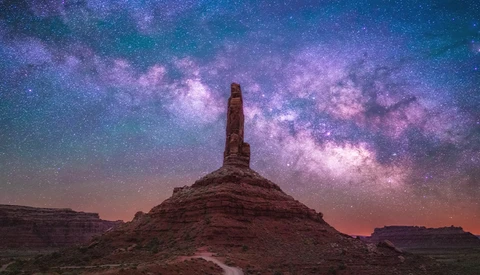 Tall rock formation silhouetted against a vibrant purple and teal Milky Way in a desert landscape.