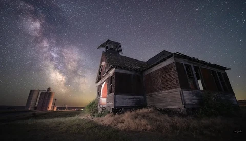 Abandoned brick schoolhouse silhouetted against the Milky Way galaxy at night.