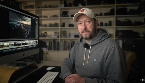 Photographer at computer workstation with editing software open and shelving with equipment in background.