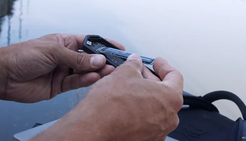 Hands holding and examining a compact camera lens or optical device against a cloudy sky backdrop.