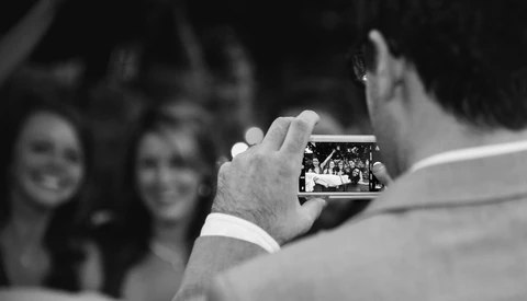 Man holding a smartphone capturing a photo of two smiling women in the background.