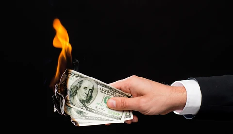 Hand in white shirt cuff holding burning U.S. dollar bills against black background.