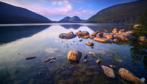 Calm alpine lake at sunrise with scattered boulders in shallow water, forested mountains in background.