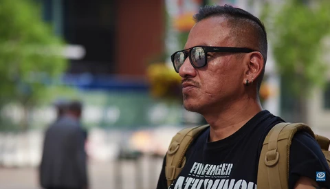 Man wearing sunglasses and tan vest standing outdoors with blurred urban background.