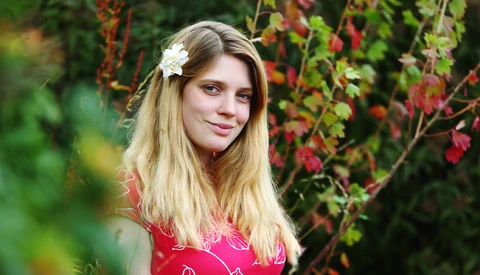 Young woman with long blonde hair and white flower accessory posing against flowering vines.