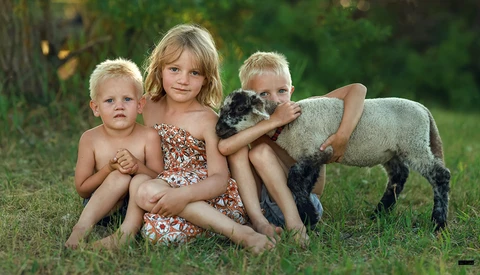 Three young children sitting on grass with a lamb, smiling at the camera.
