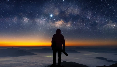 Silhouetted figure standing on a mountain peak beneath the Milky Way galaxy at twilight.