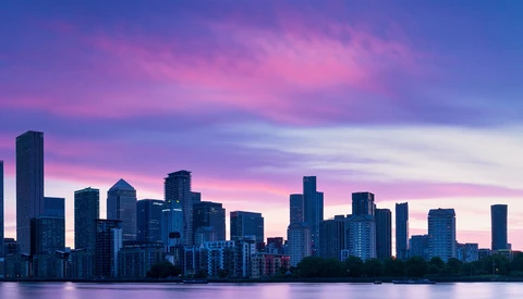 City skyline silhouetted against a vibrant purple and pink sunset sky reflected in still water below.
