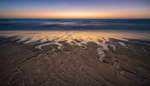 Wet sand beach at twilight with rippled patterns and gentle waves under a pastel dawn sky.