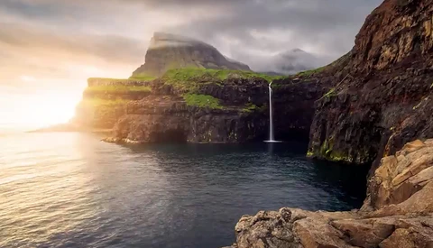 Dramatic coastal landscape with waterfall cascading into rocky cove beneath towering cliff faces at golden hour.