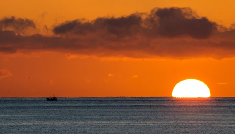 Large sun setting over calm ocean waters with a small fishing vessel silhouetted on the horizon beneath dramatic orange and brown clouds.