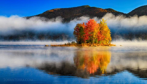 Small forested island with vibrant autumn foliage reflected in still water, surrounded by mist-covered mountains.