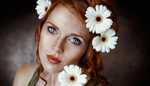 Red-haired woman with freckles and blue eyes adorned with white daisies in her hair.