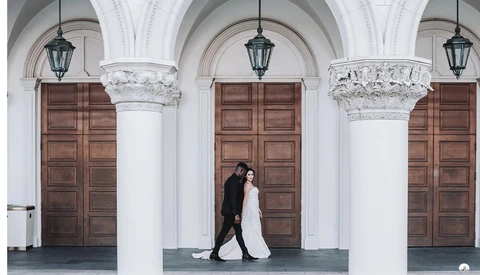 Bride and groom walking between white columns under classical arches with wooden doors.