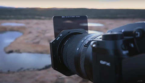 Camera with wide-angle lens mounted on tripod, pointing toward expansive landscape with distant water and hills.