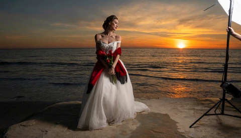 Bride in red and white off-shoulder gown holding bouquet on beach at sunset.