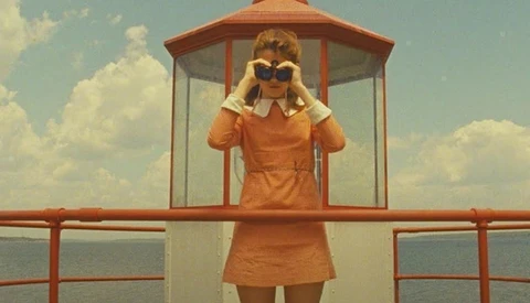 Woman in orange dress and sunglasses standing in vintage lifeguard tower overlooking calm water.