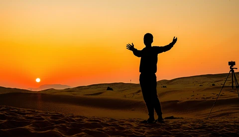 Silhouetted figure with arms raised standing in desert at sunset, with tripod-mounted camera visible on right..