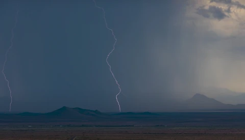 Lightning bolt streaks across stormy sky above distant mountain ranges in arid landscape.
