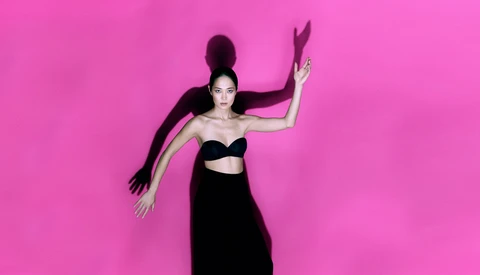 Woman in black crop top and skirt posed against bright magenta background with flowing hair and hand gestures.