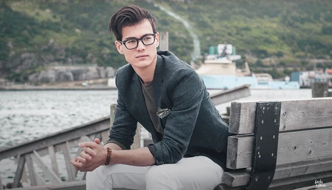Young man wearing glasses and dark jacket seated on wooden dock railing overlooking water and forested hillside.