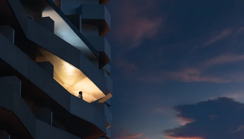 Silhouetted figure standing in an illuminated balcony alcove of a modern building at dusk.