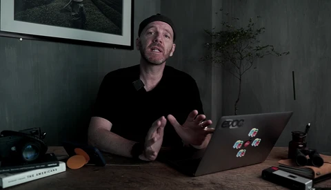 Man speaking while gesturing at laptop displaying stickers in dimly lit studio workspace.
