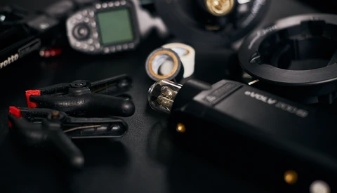 Photography and audio gear arranged on black surface including camera flash, wireless microphone, headphones, and camera body with LCD screen.