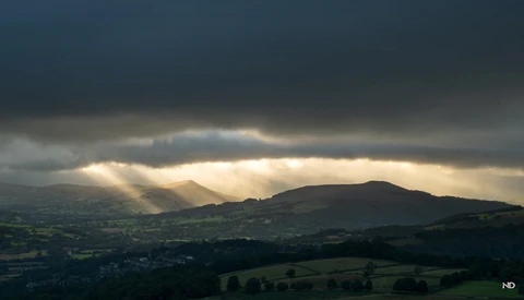 Dramatic landscape with crepuscular rays breaking through storm clouds above rolling hills and valleys.